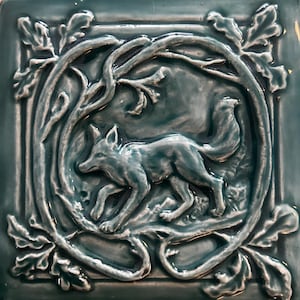 Könnte beinhalten: Eine quadratische Keramikfliese mit glänzender blaugrüner Glasur und einem Relief eines Fuchses, der durch einen kreisförmigen Rahmen aus verschlungenen Zweigen und Eichenblättern läuft. Der Fuchs ist im Profil dargestellt und von Laub umgeben.