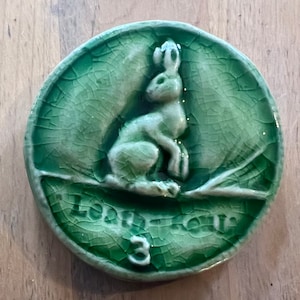 Puede incluir: Azulejo de cerámica verde con una imagen en relieve de un conejo sentado en una rama. El azulejo tiene un esmalte craquelado y el número 3 es visible en la esquina inferior derecha. Las palabras "Easter Egg" están escritas en una fuente cursiva en la parte inferior del azulejo.