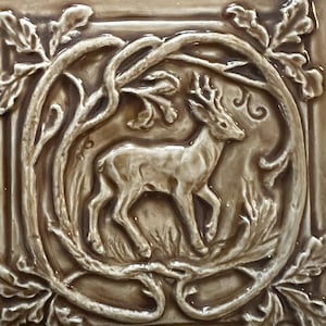 May include: A square ceramic tile with a glossy light brown finish featuring a raised relief design of a deer standing within a circular frame of intertwined branches and oak leaves. The deer is depicted in profile facing right.