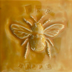 May include: A yellow ceramic tile with a white bee in relief. The bee has a white body and black stripes. The tile has the words "Bee Happy" written in white letters on it.