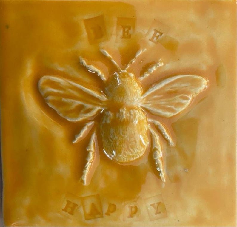 May include: A yellow ceramic tile with a white bee in relief. The bee has a white body and black stripes. The tile has the words "Bee Happy" written in white letters on it.