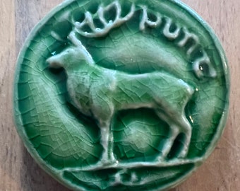 May include: A green ceramic tile with a raised image of a deer with antlers. The tile is cracked and has a rustic look. The word "PUNT" is written around the edge of the tile.
