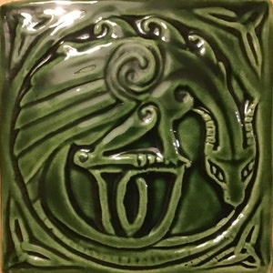 Puede incluir: Un azulejo de cerámica verde con un diseño en relieve de un dragón con un nudo celta. El dragón está enroscado alrededor de un círculo con un diseño en espiral.