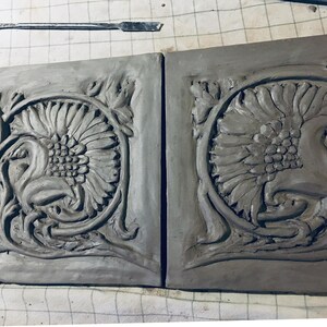 May include: Two gray clay tiles with a raised design of a stylized sunflower and floral motifs. The tiles are unfinished and have a rough texture.