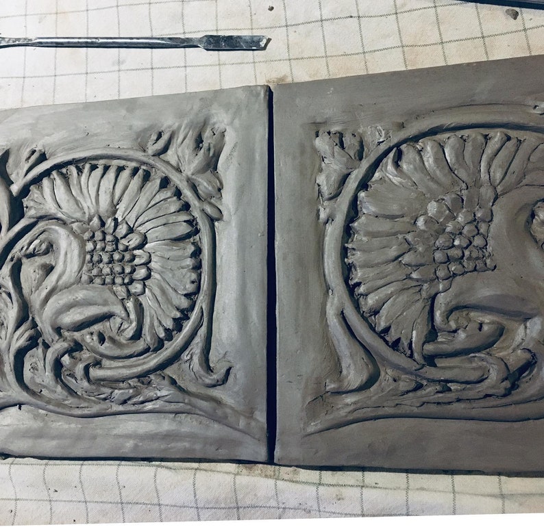 May include: Two gray clay tiles with a raised design of a stylized sunflower and floral motifs. The tiles are unfinished and have a rough texture.