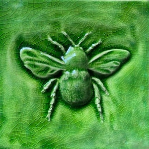 May include: A green ceramic tile featuring a raised image of a bee. The bee is green and has a textured surface.