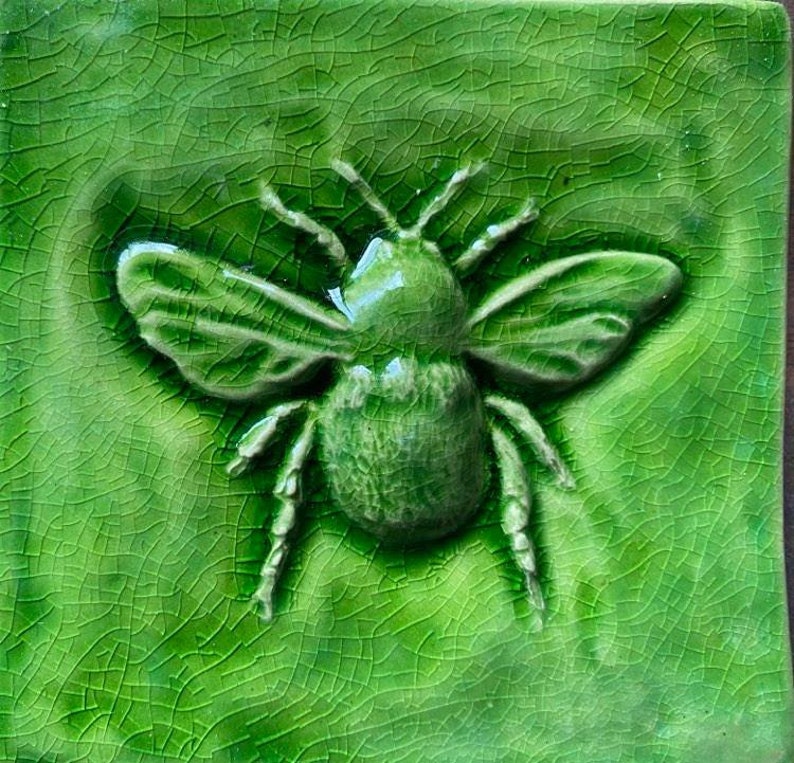 May include: A green ceramic tile with a raised image of a bee. The bee is green and has a textured surface.