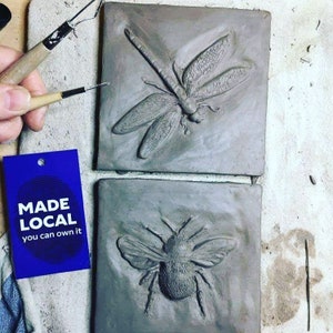 May include: Two ceramic tiles with raised clay impressions of a dragonfly and a bee. The tiles are gray and have a rough texture. A blue tag with the text "MADE LOCAL you can own it" is in the lower left corner of the image.
