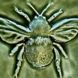 May include: A green ceramic tile with a raised image of a bee. The bee is detailed with a textured body and wings.