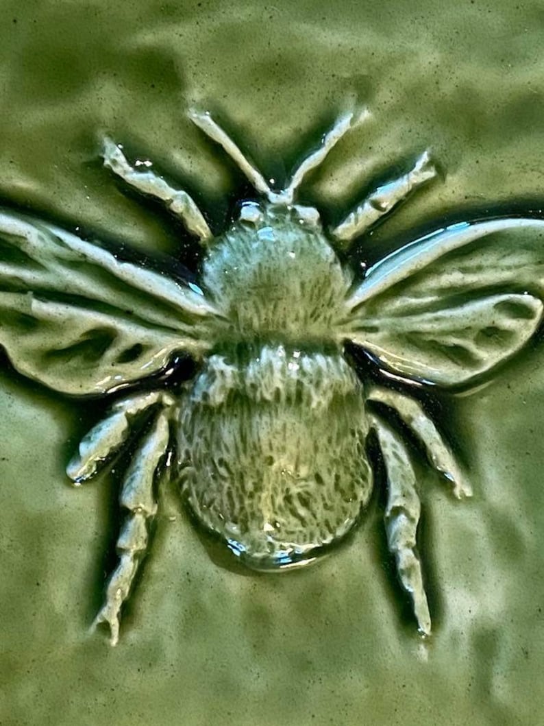 May include: A green ceramic tile with a raised image of a bee. The bee is detailed with a textured body and wings.