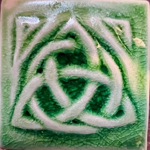 May include: Square ceramic tile with a Celtic knot design. The tile features a green background with a white, interwoven knot pattern. The glaze has a crackle effect, adding texture and visual interest to the piece.