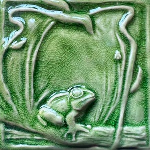 May include: Square ceramic tile featuring a relief design of a frog on a lily pad. The tile is predominantly green with a crackle glaze. The frog is light green with white detailing, and the lily pad and foliage are a darker green.