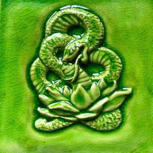 Puede incluir: Azulejo de cerámica verde con un diseño en relieve. El diseño presenta una serpiente enroscada sobre una flor de loto, ambas en tonos verdes. El azulejo tiene una superficie agrietada y texturizada.