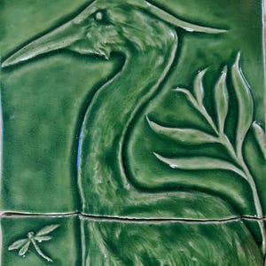 Puede incluir: Un azulejo de cerámica verde con un diseño en relieve de una garza con la cabeza girada hacia un lado. La garza está rodeada de hojas. Se puede ver una libélula en la esquina inferior izquierda del azulejo.