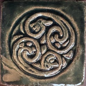 Op de afbeelding: Vierkante keramische tegel met een verhoogd Keltisch knoopontwerp in groen- en bruintinten. De tegel heeft een glanzende afwerking en een donkergroene rand, met een traditioneel Iers symbool.