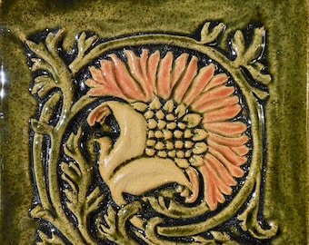 May include: A ceramic tile with a floral design. The tile is green with a brown and orange flower in the center. The flower is surrounded by a green vine.
