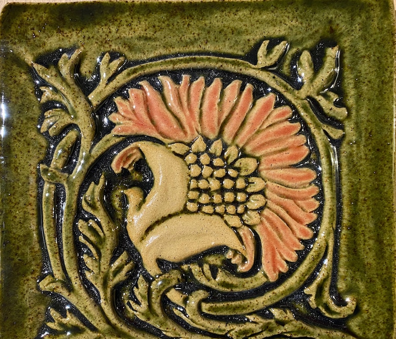 May include: A ceramic tile with a floral design. The tile is green with a brown and orange flower in the center. The flower is surrounded by a green vine.
