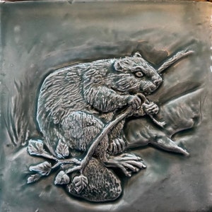 May include: A ceramic tile with a raised image of a beaver sitting on a branch. The beaver is gray and white, and the branch is brown. The tile is a light blue-green color.
