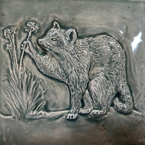 Könnte beinhalten: Eine Keramikfliese mit einer erhabenen Reliefskulptur eines Waschbären, der nach einer Pflanze greift. Die Fliese ist in einem gedämpften Grünton gehalten.