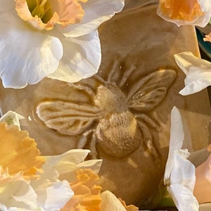 May include: A ceramic tile with a raised design of a bee, surrounded by white and orange daffodils.