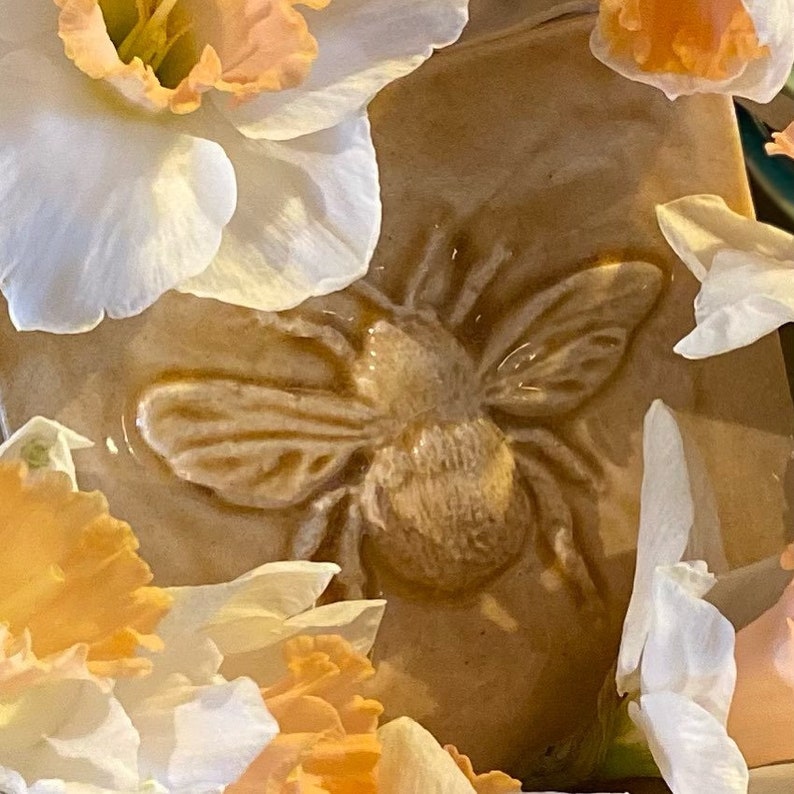 May include: A ceramic tile with a raised design of a bee, surrounded by white and orange daffodils.