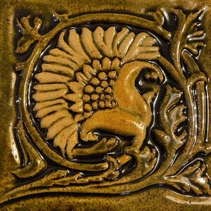May include: A brown ceramic tile with a raised design of a stylized flower with a bird perched on top. The tile has a dark brown glaze and a glossy finish.