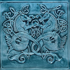 May include: Square, teal-colored ceramic tile featuring a relief design of two seahorse-like creatures facing each other. The creatures are intertwined with Celtic knotwork and other decorative elements. The tile has a glossy finish.