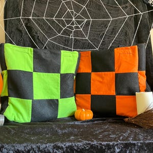 May include: Two square throw pillows with a checkerboard pattern. One pillow is green and black, the other is orange and black. A small orange pumpkin and a broom are also visible, suggesting a Halloween theme.