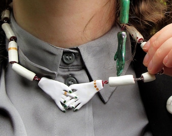 May include: A unique jewelry set featuring a necklace with white hand pendants, green and white earrings, and skull pins. The set includes rings with black and red stones. The person is wearing a gray shirt and black blazer.