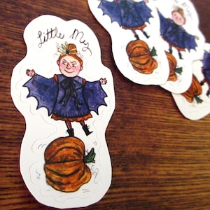 May include: A collection of whimsical stickers featuring a character named "Little My." The stickers depict a girl in a blue cape standing on a pumpkin. The text "Little My" is written above the character. The stickers are on a wooden surface.