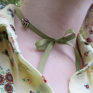 May include: A green ribbon choker necklace with a small bow and a decorative bead. The bead is white with brown and red accents. The necklace is worn with a floral print blouse.