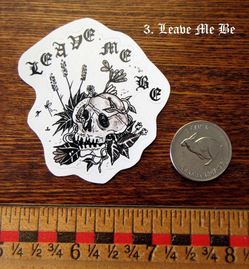 K&ouml;nnte beinhalten: Ein wei&szlig;er Aufkleber mit einem Totenkopf-Design, verziert mit floralen Elementen und dem Schriftzug "LEAVE ME BE" in gotischer Schrift. Eine kanadische 5-Cent-M&uuml;nze dient als Gr&ouml;&szlig;envergleich. Der Aufkleber liegt auf einer Holzoberfl&auml;che.