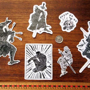 K&ouml;nnte beinhalten: Eine Sammlung von schwarz-wei&szlig;en Aufklebern mit Fantasy-Kunstmotiven. Die Aufkleber zeigen Illustrationen von Figuren mit Schwertern, Skeletten und einem Sch&auml;del mit dem Text "leave me". Ein Lineal und eine M&uuml;nze sind ebenfalls vorhanden.