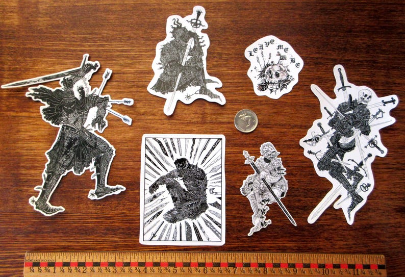 K&ouml;nnte beinhalten: Eine Sammlung von schwarz-wei&szlig;en Aufklebern mit Fantasy-Kunstmotiven. Die Aufkleber zeigen Illustrationen von Figuren mit Schwertern, Skeletten und einem Sch&auml;del mit dem Text "leave me". Ein Lineal und eine M&uuml;nze sind ebenfalls vorhanden.