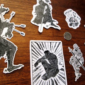 K&ouml;nnte beinhalten: Eine Sammlung von schwarz-wei&szlig;en Aufklebern mit detaillierten Illustrationen von Kriegern, Sch&auml;deln und anderen Fantasy-Motiven. Die Aufkleber sind auf einer Holzoberfl&auml;che angeordnet, eine M&uuml;nze ist sichtbar.