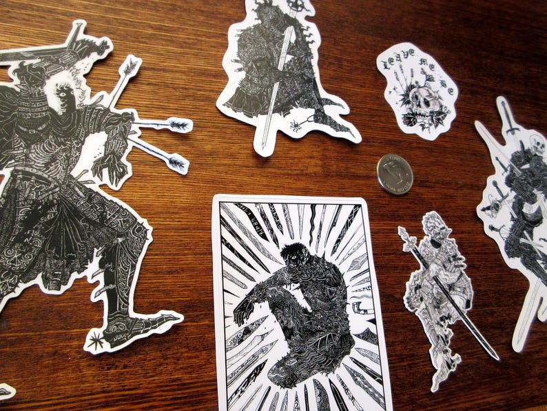 K&ouml;nnte beinhalten: Eine Sammlung von schwarz-wei&szlig;en Aufklebern mit detaillierten Illustrationen von Kriegern, Sch&auml;deln und anderen Fantasy-Motiven. Die Aufkleber sind auf einer Holzoberfl&auml;che angeordnet, eine M&uuml;nze ist sichtbar.