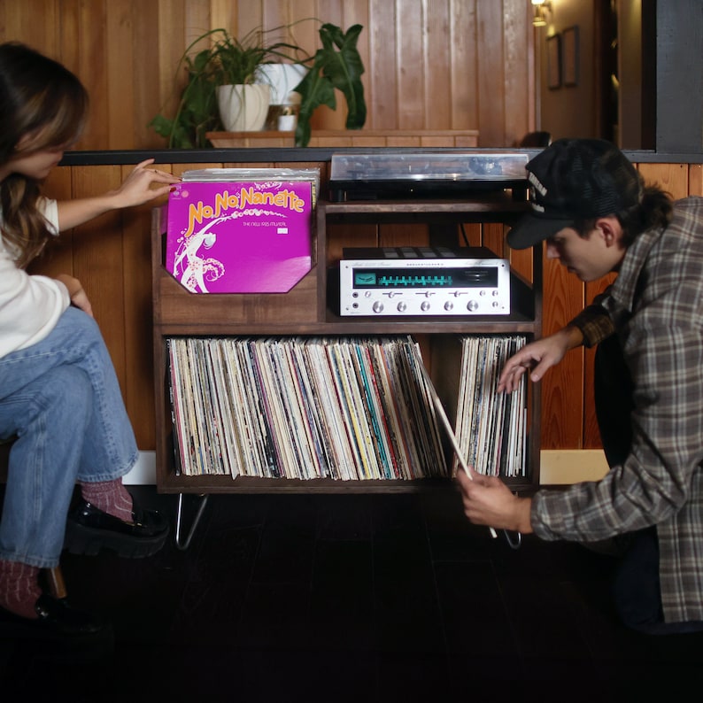 May include: A wooden record storage unit with a turntable and stereo receiver. The unit holds numerous vinyl records, with a pink album cover visible. Two people are interacting with the records.