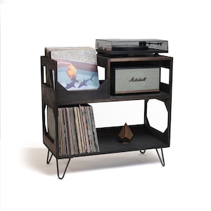May include: A dark stained wooden record player stand with a turntable, speaker, and vinyl records. The stand has three shelves, with a speaker on the middle shelf and records on the top and bottom shelves. The stand has black metal hairpin legs.