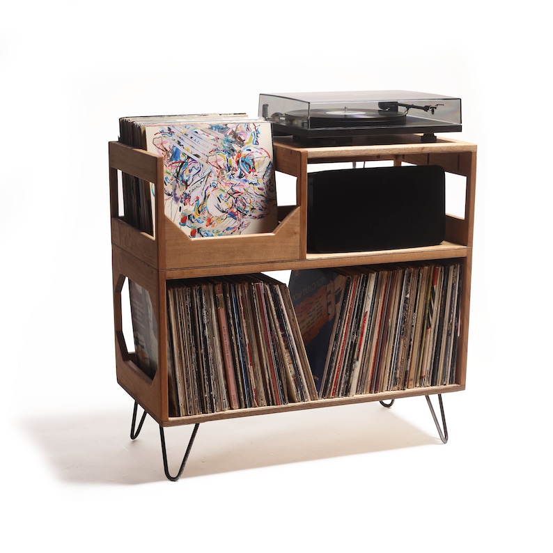 May include: A wooden record storage unit with a turntable on top. The unit has two shelves for vinyl records and a compartment for a speaker. The unit is supported by black hairpin legs. The records are in various colors.