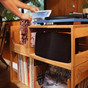 May include: A wooden record storage unit with a blue turntable on top and a black speaker below. The unit is filled with vinyl records. A person is seen placing a record on the turntable.