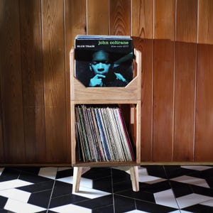 May include: Wooden record storage unit with two shelves, holding vinyl records. The top shelf displays a John Coltrane album, "Blue Train." The unit has a natural wood finish and is set against a wood-paneled wall and a black and white tiled floor.
