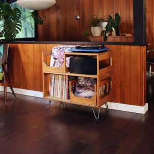 May include: A wooden record player stand with a blue turntable, black speaker, and storage for vinyl records. The stand has a mid-century modern design with metal hairpin legs. The room has wood paneling and a large window.