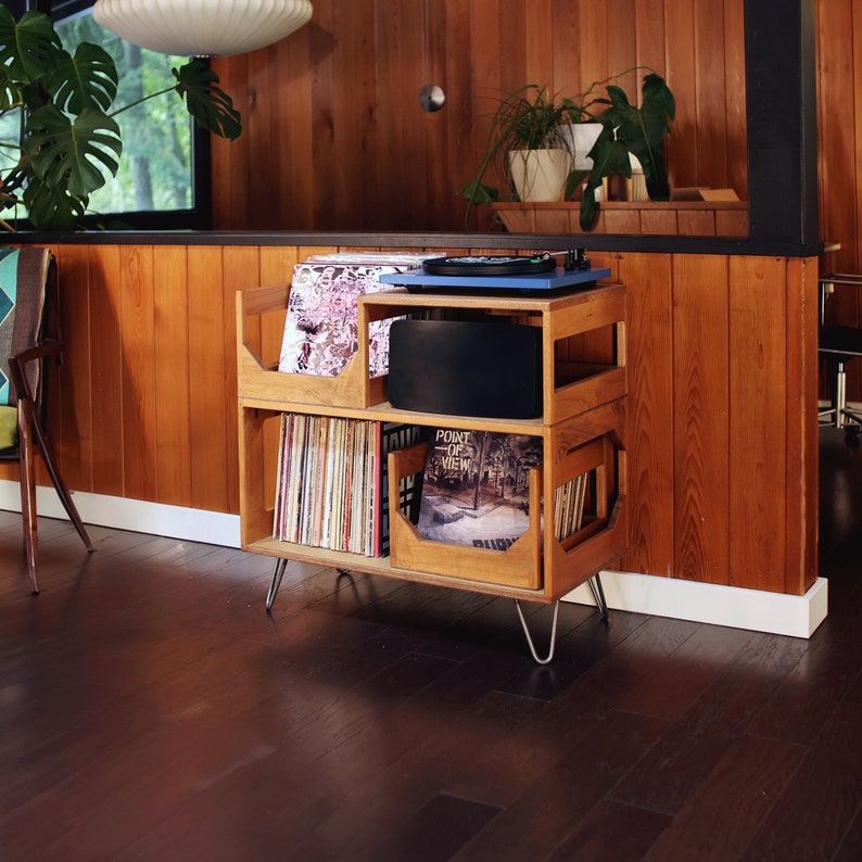 May include: A wooden record player stand with a blue turntable, black speaker, and storage for vinyl records. The stand has a mid-century modern design with metal hairpin legs. The room has wood paneling and a large window.