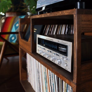 May include: A wooden record storage unit with a silver stereo receiver and a black turntable. The unit has multiple shelves filled with vinyl records. The receiver has several knobs and a blue display. The overall aesthetic is vintage and retro.