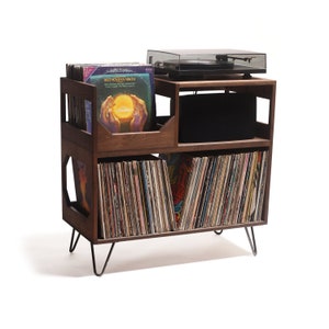 May include: A wooden record player stand with a turntable on top and storage for vinyl records. The stand is dark brown with black metal legs. The stand has multiple compartments for record storage and a shelf for the turntable.