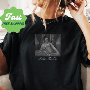 May include: A black short-sleeved t-shirt featuring a rectangular black and white graphic print of a person reclining on a bed. Below the image, the text reads I Am The Tea in a white serif font. The shirt has a crew neckline and a relaxed fit.