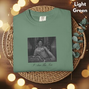 May include: A light green t-shirt featuring a black and white rectangular graphic of a person sitting in a room, with the text "I Am The Tea" printed in a cursive font below the image. The shirt has a crew neckline and a visible size label.