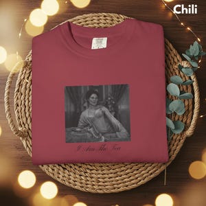 May include: A dark red crewneck t-shirt featuring a black and white rectangular graphic of a person in a vintage-style outfit. Below the graphic, the text reads I Am The Tea in a black script font. The shirt has a white interior neck label.