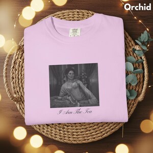 May include: A light pink t-shirt featuring a black and white rectangular graphic of a person sitting in a room. Below the graphic, the text reads I Am The Tea in a cursive font. The shirt has a crew neckline and a Comfort Colors brand label.