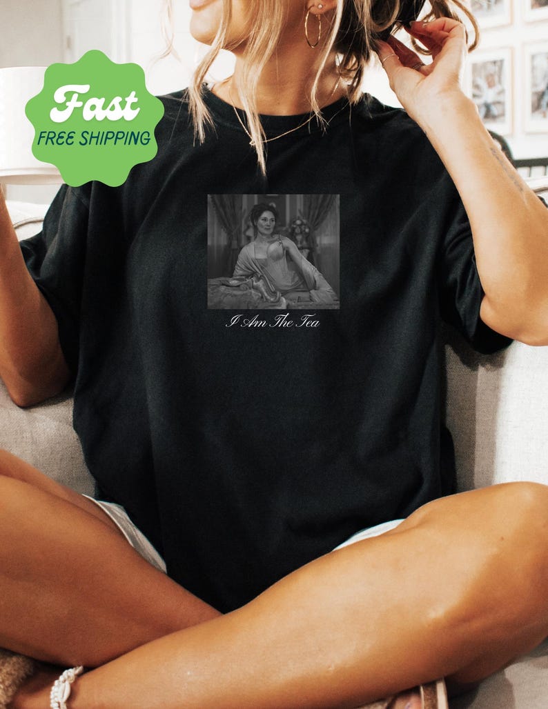 May include: A black short-sleeved t-shirt featuring a rectangular black and white graphic print of a person reclining on a bed. Below the image, the text reads I Am The Tea in a white serif font. The shirt has a crew neckline and a relaxed fit.